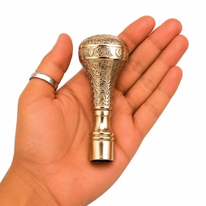 Op de afbeelding: Een gepolijste messing handgreep met een afgeronde bovenkant en een taps toelopende schacht, voorzien van ingewikkelde bloemengravures op het bovenste gedeelte en een gladde, cilindrische basis. Het metalen oppervlak reflecteert het licht.