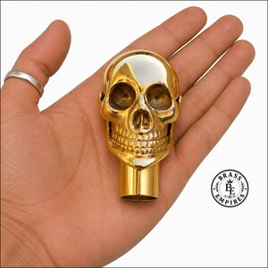 Puede incluir: Un mango o remate en forma de calavera de latón pulido de color dorado con cuencas oculares profundas, una cavidad nasal definida y dientes detallados. Un punto de fijación cilíndrico se extiende desde la base. El logotipo Brass Empires es visible en la esquina.