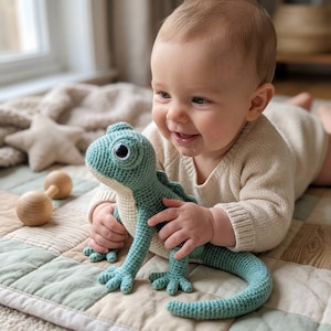 Puede incluir: Un peluche de lagarto tejido a ganchillo en colores verde azulado y crema, con grandes ojos de plástico en blanco y negro, un cuerpo texturizado y una cola larga. El juguete presenta intrincadas puntadas de hilo en toda su forma, mostrando un diseño reptiliano detallado.