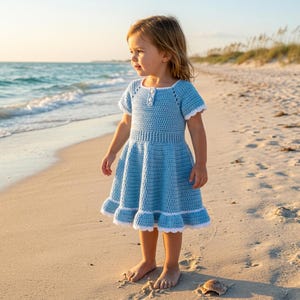 Pode incluir: Vestido de crochê azul claro com acabamento branco. O vestido apresenta mangas curtas, corpete justo e saia rodada. Uma criança usa o vestido em uma praia de areia perto do oceano.