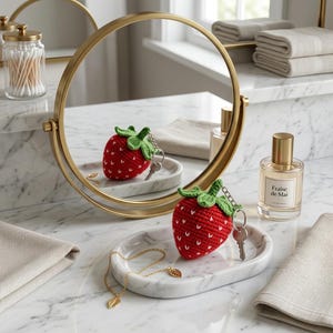 May include: A red crocheted strawberry keyring with a green stem and white embroidered seeds, resting on an oval white marble tray alongside a delicate gold chain necklace with a leaf charm, positioned in front of a round gold-framed mirror.
