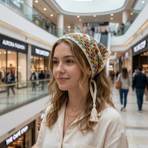 Puede incluir: Un pañuelo triangular de ganchillo con un patrón de cuadrados de la abuela multicolor en tonos azul, verde, amarillo y óxido, acabado con un borde color crema y lazos trenzados con borlas que se aseguran debajo del cabello.
