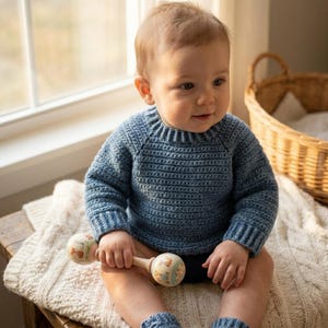 May include: A baby wearing a blue crochet sweater and matching leg warmers sits on a wooden bench. The baby holds a wooden rattle with painted animal designs. A woven basket and a cream-colored blanket are in the background.
