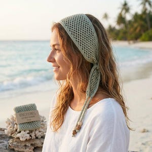 Pode incluir: Uma faixa de cabelo de crochê verde claro com um padrão de losangos, amarrada nas costas. A faixa de cabelo é usada por uma pessoa com cabelo loiro longo e ondulado. O texto "VIVCROCHETS" é visível em uma embalagem pequena. O fundo é uma praia com palmeiras e oceano.