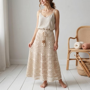 May include: A cream-coloured, open-weave crochet skirt with a drawstring waist and wooden bead accents. The skirt is paired with a neutral-toned camisole. A wicker chair with yarn and crochet hooks is in the background.