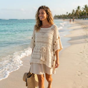 May include: Cream-colored crochet top with short sleeves and wooden buttons. The top has a drawstring waist and is paired with a beige skirt. A straw tote bag and hat are also visible. The outfit is perfect for a beach day.