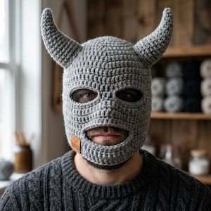 Könnte beinhalten: Eine graue gehäkelte Sturmhaube mit zwei gebogenen Hörnern. Die Maske hat Augen- und Mundöffnungen, und ein kleines braunes Etikett ist angebracht. Die Person im Bild trägt einen grauen Pullover.