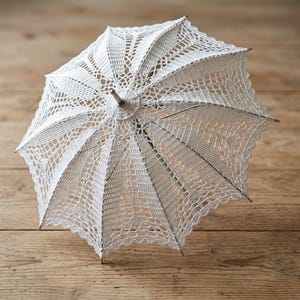 Op de afbeelding: Een witte, opengewerkte gehaakte parasol met een zilverkleurig handvat en frame. Het ingewikkelde kanten ontwerp creëert een delicate, luchtige uitstraling. De parasol ligt op een houten oppervlak.