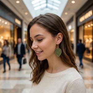 May include: A close-up of a woman wearing a pair of green leaf-shaped crochet earrings. The earrings are suspended from gold-colored hooks. The woman is wearing a cream-colored sweater.