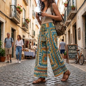 May include: A person wearing colorful, wide-leg crochet pants with a drawstring waist and tassels. The pants feature a geometric pattern in shades of blue, yellow, and orange. The person is also wearing brown sandals.