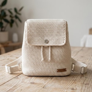 Pode incluir: Uma mochila de crochê creme com fecho de aba e alças ajustáveis. A bolsa possui um fecho de cordão e uma pequena etiqueta retangular com texto. A mochila está sobre uma superfície de madeira.