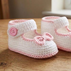May include: A pair of white crochet baby shoes with pink trim and a pink bow. One shoe features a pink button fastening. The shoes are designed for infants and are handmade.