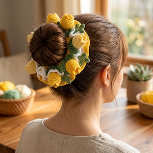 Puede incluir: Un accesorio para el cabello de ganchillo amarillo, adornado con flores amarillas en forma de tulipán y hojas verdes, se usa en un moño. El accesorio se coloca sobre un fondo de cabello castaño y una mesa de madera. El tema general son los accesorios para el cabello hechos a mano.