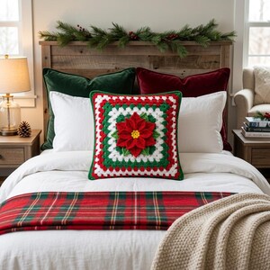 Könnte beinhalten: Ein quadratisches, gehäkeltes Kissen mit einem Poinsettien-Design in Rot, Grün und Weiß. Das Kissen liegt auf einem Bett mit weißer Bettwäsche, einem rot-grünen karierten Läufer und grünen und roten Samtkissen. Im Hintergrund befindet sich ein hölzernes Kopfende.