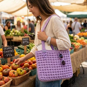 Pode incluir: Uma sacola tote de crochê lilás com um padrão de ponto waffle. A sacola tem formato retangular e uma alça combinando. Óculos de sol estão presos à sacola. A sacola está sendo segurada em uma feira com tomates e outros produtos.