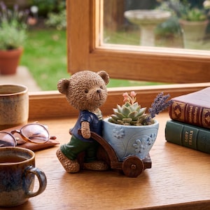 Puede incluir: Una figura de oso de peluche marrón empujando un carrito de maceta azul lleno de una suculenta y lavanda. La maceta tiene un diseño floral. El oso lleva una camisa azul y pantalones verdes. La escena está ambientada sobre una superficie de madera cerca de una ventana.
