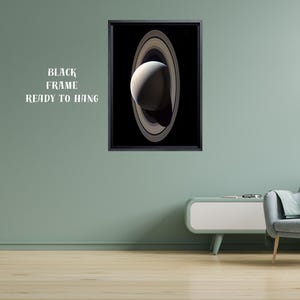 May include: A framed wall art print featuring a vertical depiction of the planet Saturn with its prominent rings against a solid black background. The artwork is enclosed in a thin black frame and includes the text BLACK FRAME READY TO HANG.