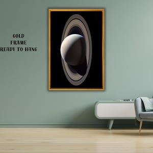 May include: A rectangular wall art piece featuring a high-contrast image of a planet with prominent rings set against a black background, enclosed in a thin gold frame. The text GOLD FRAME READY TO HANG is printed in black on the wall nearby.