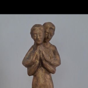 May include: A gold-toned sculpture depicting two figures standing together with their hands pressed together in a prayer-like gesture. The figures are joined at the back, sharing a single base, and feature a textured, metallic finish throughout.