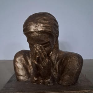 May include: A bronze-colored sculpture of a person from the chest up, with their head bowed and both hands covering their face. The figure has a textured surface and is mounted on a rectangular base, capturing a pose of deep contemplation or sorrow.
