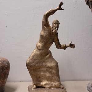 May include: A gold-toned sculpture of a figure in a long, textured robe standing on a rectangular base. The figure is depicted in a dynamic pose with one arm raised upward and the other extended outward, featuring a rough, weathered surface texture.