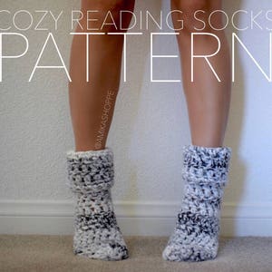 May include: A pair of cozy crochet socks in black and white. The socks are worn on a person's feet. The text "Cozy Reading Socks Pattern" is at the top of the image. The text "@amikashoppe" is on the side of the image.