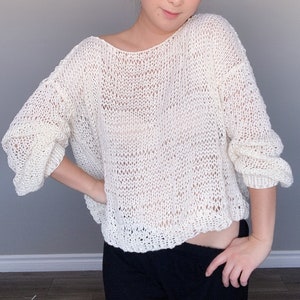 Könnte beinhalten: Ein locker sitzender, weißer Strickpullover mit einem weiten, offenen Ausschnitt. Der Pullover hat ein zartes, offenes Strickmuster und eine leicht verkürzte Länge.