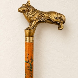 Könnte beinhalten: Ein Gehstock mit einem goldfarbenen Metallgriff in Form eines stehenden Hundes. Der Schaft besteht aus orangefarbenem Holz mit einem dunklen, astartigen Muster und feinen türkisfarbenen Adern, die sich über die gesamte Länge des Holzes ziehen.