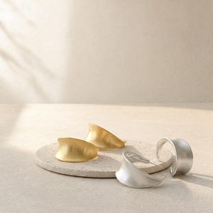 May include: Two pairs of hoop earrings with a wide, curved, and flared silhouette. One pair is finished in a matte gold tone, while the other pair features a matte silver tone. The earrings have a smooth, solid surface and a distinct concave shape.