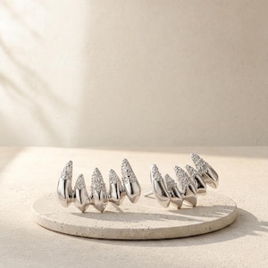 May include: A pair of silver-toned ear climbers featuring a multi-pronged claw design. Each piece consists of five curved, pointed segments, with the upper portions of each prong encrusted with small, clear, sparkling stones in a pavé setting.