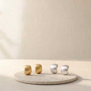 May include: Two pairs of chunky hoop earrings, one pair in a matte gold finish and one pair in a matte silver finish. The earrings have a smooth, curved, and thick design, positioned upright on a circular, textured, light-colored stone base.