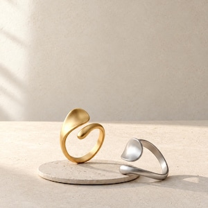 May include: Two open-style rings with smooth, rounded, organic shapes. One ring is finished in a matte gold tone, while the other is finished in a matte silver tone. Both rings feature a curved, asymmetrical design with a gap between the ends.