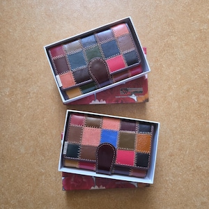 Peut inclure: Deux portefeuilles en cuir avec un motif patchwork multicolore, comprenant du rouge, du bleu, du marron et du vert. Chaque portefeuille est dans une boîte blanche avec un motif floral.