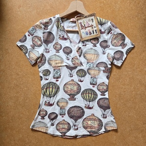 Puede incluir: Una camiseta blanca de manga corta con cuello en V y un estampado integral de globos aerostáticos de estilo vintage en tonos marrón, beige y dorado. Una etiqueta de cartón rectangular con el texto "Leyenda & Co" está unida al cuello.