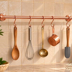 Puede incluir: Una barra de cocina de metal color cobre montada horizontalmente con cinco ganchos en S a juego. Colgados de los ganchos hay una cuchara de madera, un batidor de metal, un cucharón color latón, una taza medidora de cobre y una espátula de silicona gris.