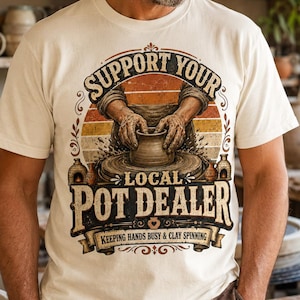 May include: A cream-colored short-sleeve t-shirt featuring a graphic of hands shaping clay on a pottery wheel. The text reads "SUPPORT YOUR LOCAL POT DEALER" and "KEEPING HANDS BUSY & CLAY SPINNING" in a vintage-style font with orange accents.