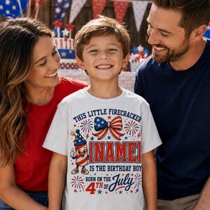 Puede incluir: Una camiseta gris claro de manga corta con un gráfico en rojo, blanco y azul que muestra un petardo de dibujos animados, un lazo y el texto: This Little Firecracker [NAME] Is The Birthday Boy Born On The 4th Of July, decorado con motivos de estrellas y fuegos artificiales.