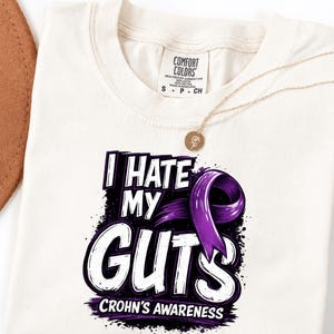 Op de afbeelding: Crèmekleurig T-shirt met de tekst "I HATE MY GUTS" in wit en paars, met een paars lint en de woorden "CROHN'S AWARENESS" eronder. Een gouden ketting met een kleine hanger is over het shirt gedrapeerd.