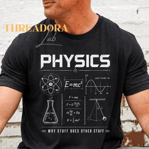 Puede incluir: Camiseta gris oscuro con la palabra "PHYSICS" en blanco. La camiseta presenta diagramas científicos, fórmulas y la frase "WHY STUFF DOES OTHER STUFF" en blanco. También se ve un gráfico de un átomo, un matraz y un péndulo.