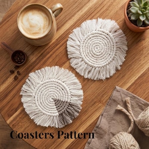 May include: Two off-white, circular macrame coasters with a spiral design and fringe detail. The coasters are on a wooden table, alongside a cup of coffee, a small succulent plant, and the text "Coasters Pattern".