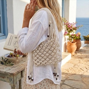 Può includere: Una borsa a tracolla in macramè color crema con lunghe frange e perline di legno. La borsa presenta un motivo intrecciato geometrico. La borsa è indossata sulla spalla. Un libro con testo è tenuto in mano.