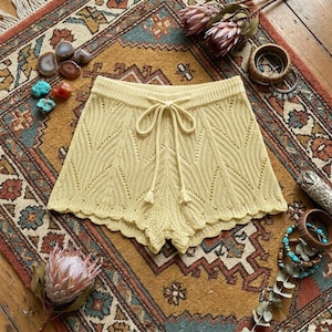 May include: Pale yellow knit shorts with a scalloped hem and drawstring waist. The shorts feature a textured, openwork design. The garment is displayed on a patterned rug with decorative items.
