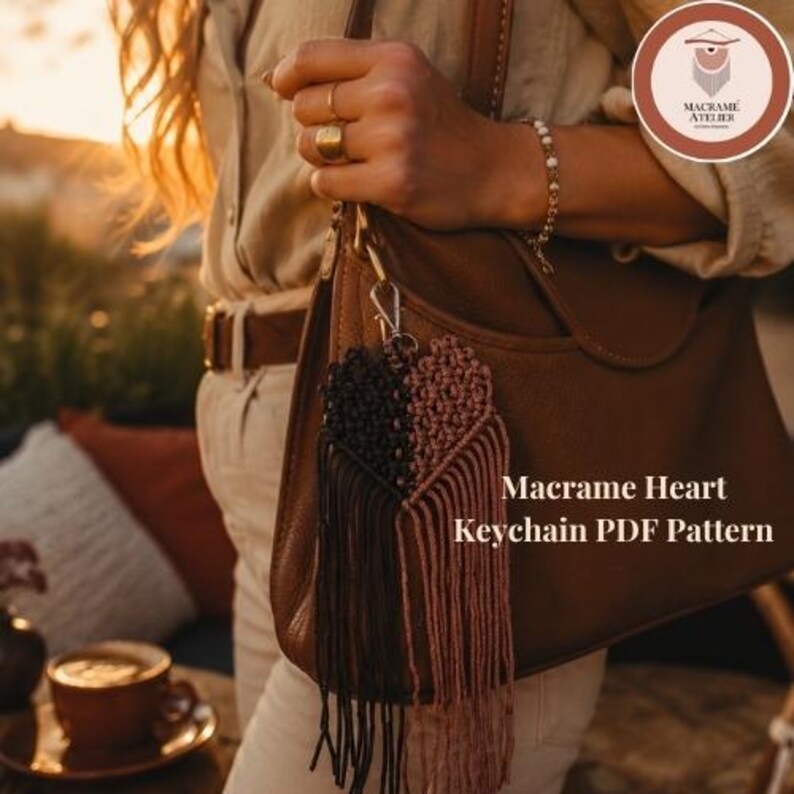 Puede incluir: Un llavero de coraz&oacute;n de macram&eacute; con dos mitades en cord&oacute;n negro y rosa polvoriento, sujeto a un bolso de cuero marr&oacute;n con un cierre de metal plateado. El texto Macrame Heart Keychain PDF Pattern aparece impreso a la derecha.