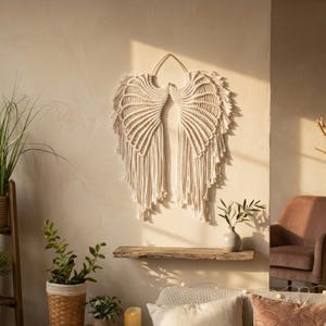 May include: A cream-colored macrame wall hanging shaped like a pair of angel wings. The piece is constructed from knotted cotton cord, featuring intricate fan-like patterns that extend into long, loose fringe strands hanging vertically downward.
