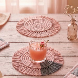 May include: Two round macrame coasters featuring a spiral cord design with fringed edges. One coaster is solid light pink, while the other is light pink with a segment of dark grey fringe. A glass of pink liquid sits on the multi-coloured coaster.