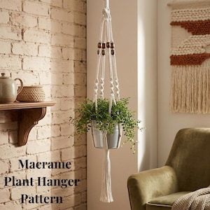 May include: A white macrame plant hanger with a metal pot holding a green plant. The macrame design includes wooden beads and a tassel. Text on the image reads "Macrame Plant Hanger Pattern".