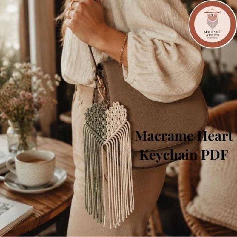 Puede incluir: Un llavero de coraz&oacute;n de macram&eacute; con un dise&ntilde;o dividido, una mitad en verde salvia y la otra en crema, terminado con largos cordones de flecos colgantes. El llavero est&aacute; sujeto a un bolso de hombro marr&oacute;n con un cierre de metal dorado.