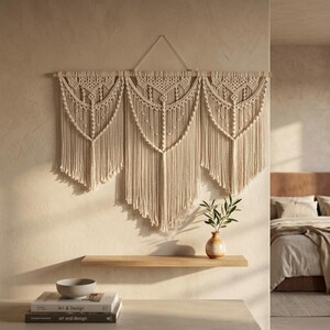 Può includere: Grande arazzo da parete in macramè color crema. Il design presenta tre pannelli collegati con nodi intricati e frange. L'arredamento murale è montato su un tassello di legno, aggiungendo un tocco naturale allo stile bohémien.