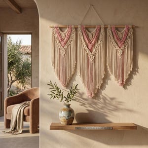 Könnte beinhalten: Drei Makramee-Wandbehänge in Creme- und Rosatönen. Jedes Stück hat ein geometrisches Design und lange Fransen, die an einer Holzstange aufgehängt sind. Die Breite beträgt 110 cm, die mittlere Länge 62 cm und die Seitenlänge 85 cm.