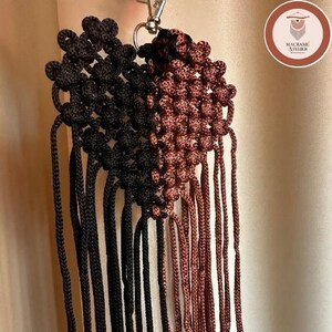 Puede incluir: Un llavero de macram&eacute; en forma de coraz&oacute;n con un dise&ntilde;o dividido, con cord&oacute;n negro a la izquierda y cord&oacute;n marr&oacute;n a la derecha. La pieza est&aacute; construida con patrones de nudos intrincados e incluye largos flecos colgantes unidos a un cierre de metal plateado.
